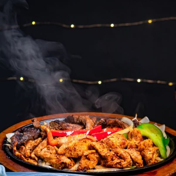 Sizzling chicken and steak fajitas with onions and peppers on a hot plate, emitting steam.