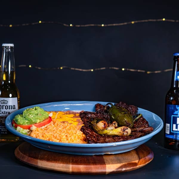 El Mirador 2 carne asada with rice, beans, guacamole, and two beer bottles on a dark background.