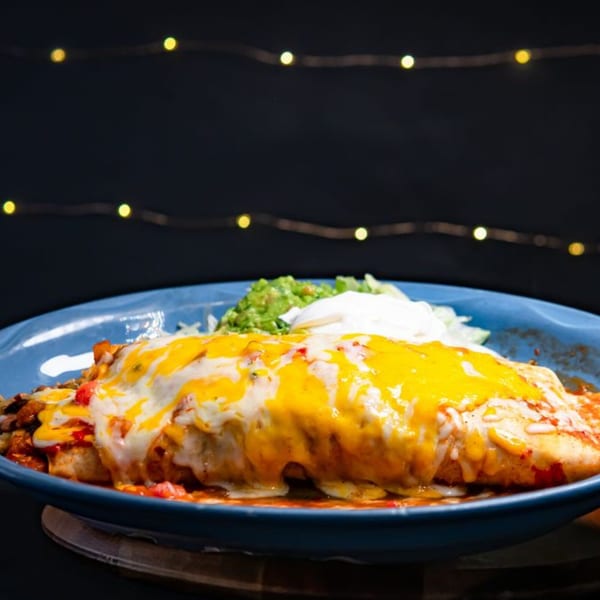 A large, cheesy burrito with guacamole and sour cream on a blue plate, against a dark background with string lights.