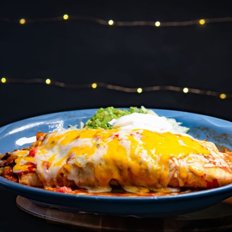 A large, cheesy burrito with guacamole and sour cream on a blue plate, against a dark background with string lights.